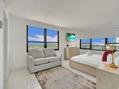 a white bedroom with a bed and a chair and windows at Beautiful Beach Condo Beach Service 1020 in Miami Beach