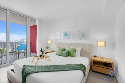 a white bedroom with a large bed and a balcony at Modern Resort Retreat Steps from the Beach in Hallandale Beach