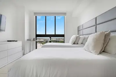 a white bedroom with two beds and a window at La Central- CityPlace Doral and across from a Park in Miami