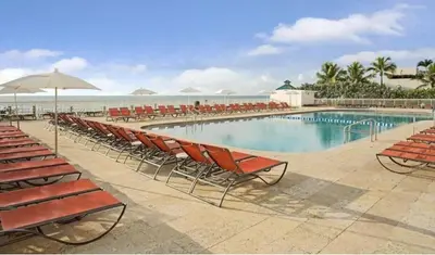 a pool with chairs and an umbrella and the ocean at Ramada Marco Polo Beach in Miami Beach